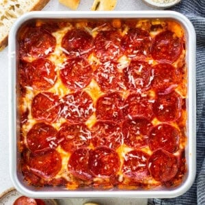 Square deep-dish pepperoni pizza in a pan, surrounded by bread slices, grated cheese in a bowl, and a small plate of pepperoni slices on a light surface.