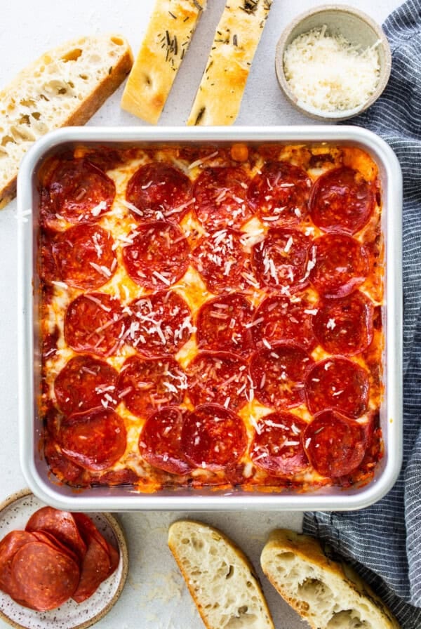 Square deep-dish pepperoni pizza in a pan, surrounded by bread slices, grated cheese in a bowl, and a small plate of pepperoni slices on a light surface.