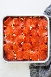 Square baking dish with layered pepperoni slices on top of shredded cheese and pasta. Gray cloth partially visible at the bottom right.