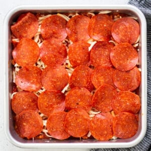 Square baking dish with layered pepperoni slices on top of shredded cheese and pasta. Gray cloth partially visible at the bottom right.