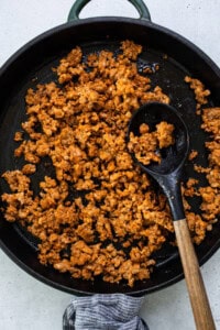 Cooked crumbled sausage in a black skillet with a wooden spoon on a light surface.