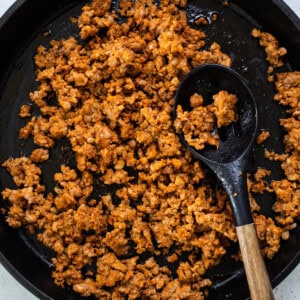 Cooked crumbled sausage in a black skillet with a wooden spoon on a light surface.