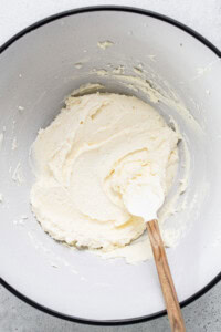 Mixing bowl with creamy, light-colored batter being stirred by a white spatula with a wooden handle.