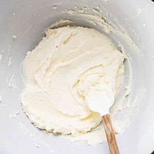 Mixing bowl with creamy, light-colored batter being stirred by a white spatula with a wooden handle.