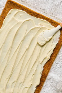 A rectangular cake layer being spread with cream cheese frosting using a spatula on a floured surface.