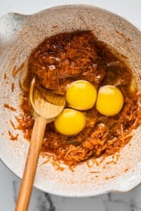 Three raw eggs are cracked into a mixing bowl containing brown shredded ingredients, with a wooden spoon partially submerged in the mixture.