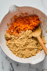 A mixing bowl containing shredded carrots and brown sugar, with a wooden spoon resting inside.