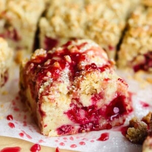 A close-up of a slice of crumb cake with a raspberry filling and a drizzle of raspberry sauce on top. Dustings of powdered sugar are visible on the surface.