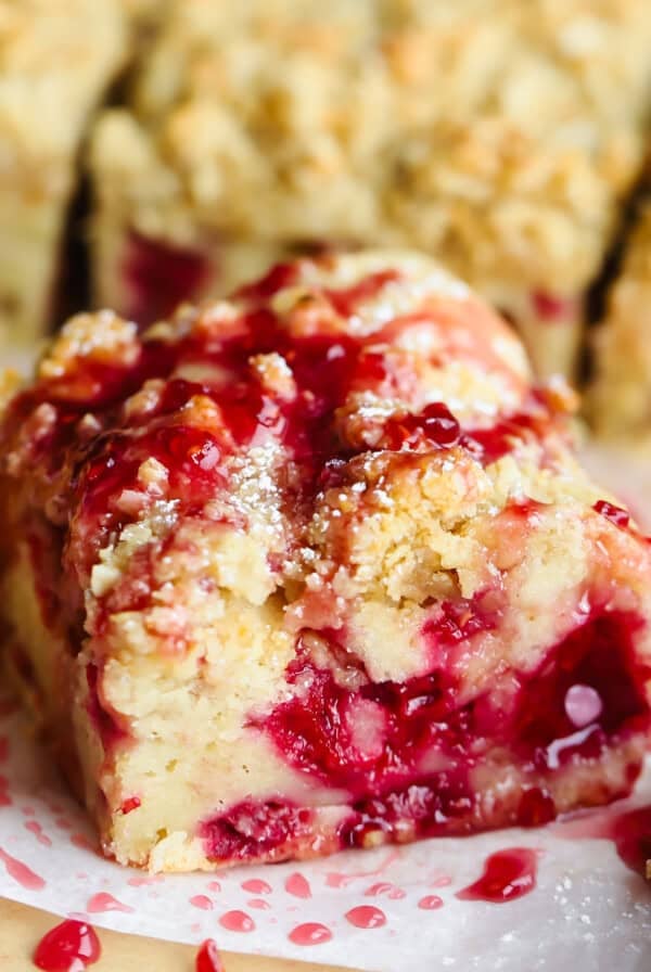 A close-up of a slice of crumb cake with a raspberry filling and a drizzle of raspberry sauce on top. Dustings of powdered sugar are visible on the surface.