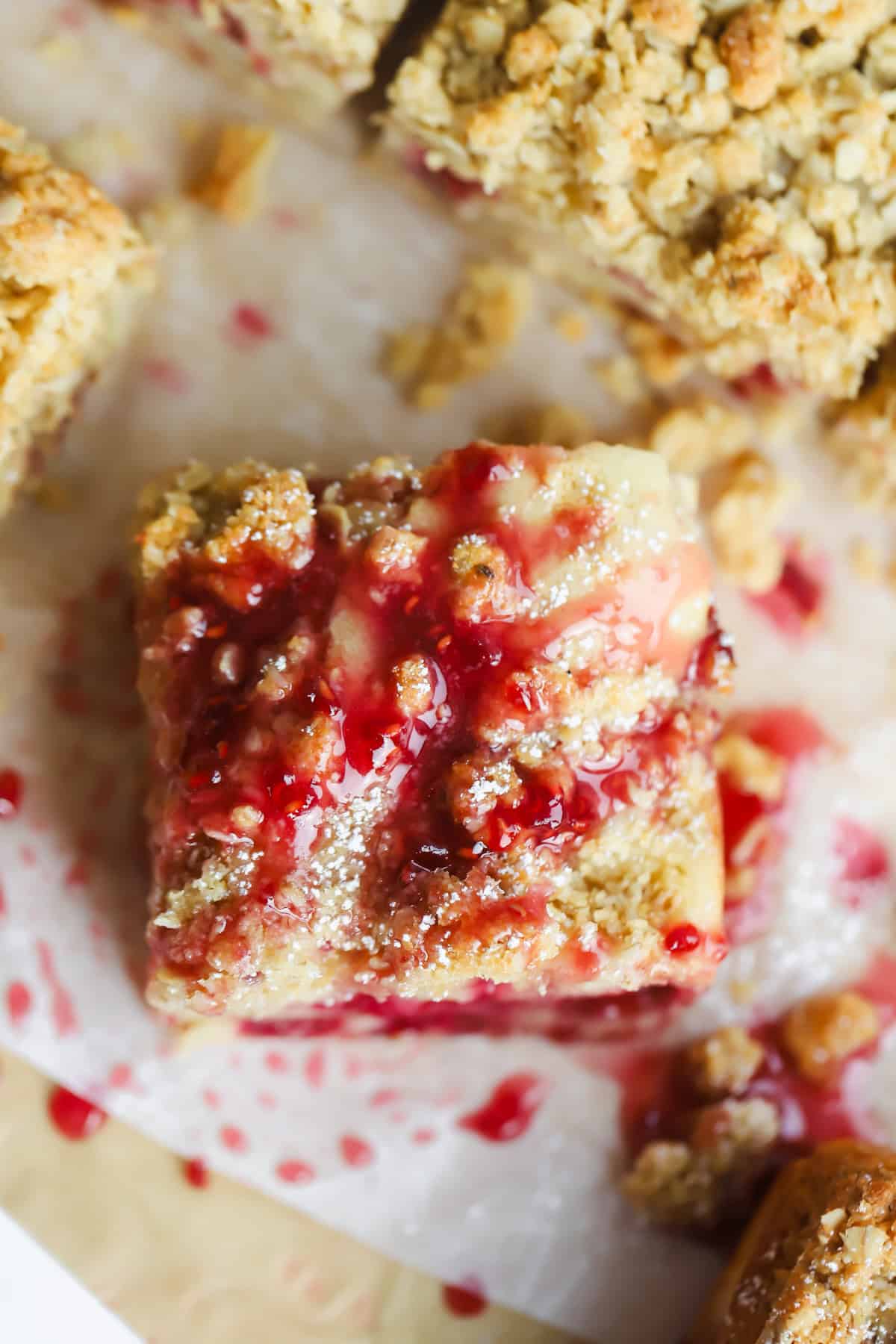 A square piece of dessert topped with a crumbly streusel and drizzled with red fruit sauce on parchment paper.