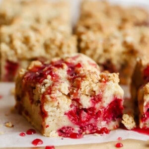 Close-up of crumbly dessert bars with a raspberry filling, topped with a drizzle of red sauce, arranged on parchment paper.