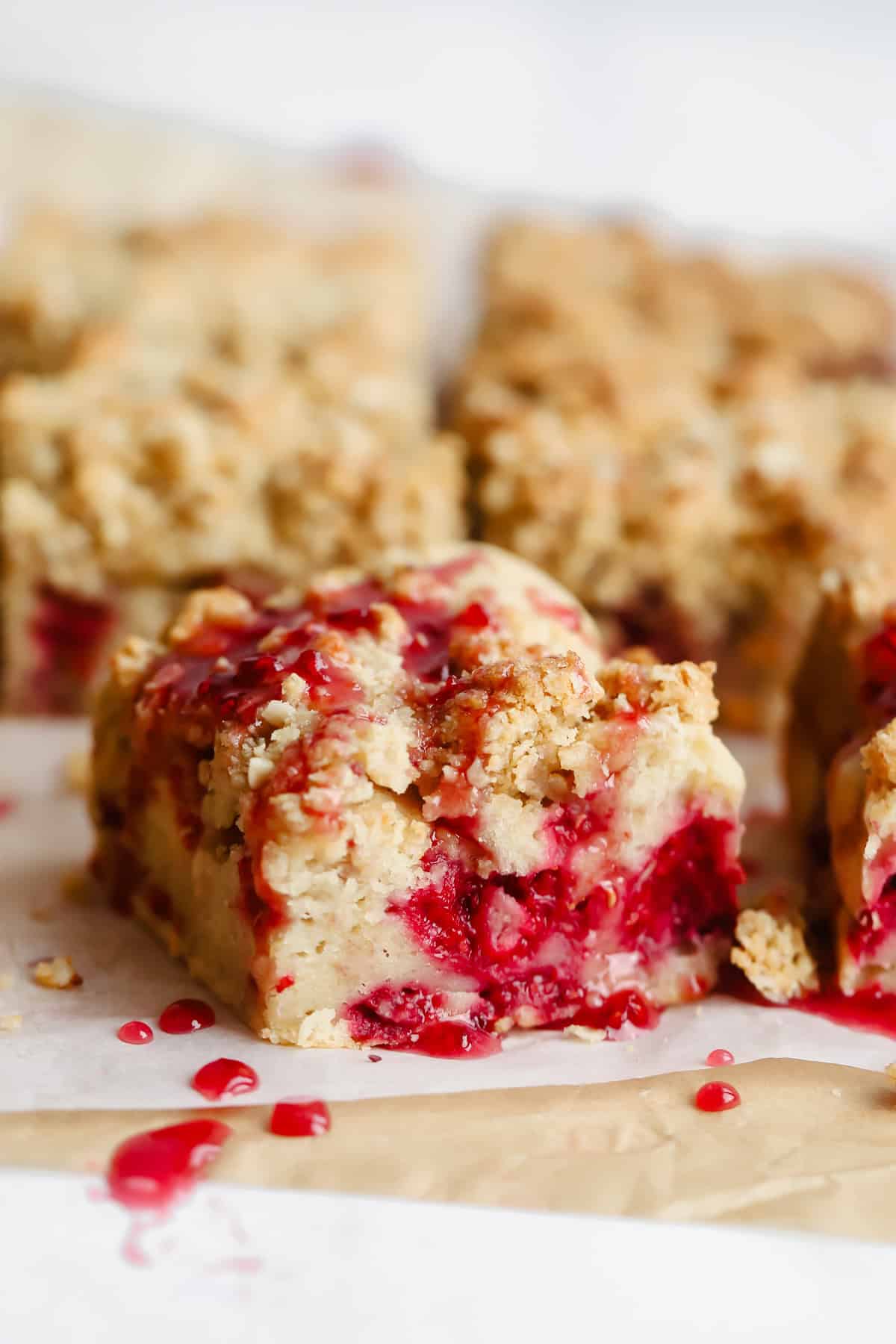 Close-up of crumbly dessert bars with a raspberry filling, topped with a drizzle of red sauce, arranged on parchment paper.