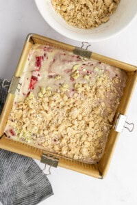 Square baking pan with strawberry batter topped with crumble next to a bowl of crumble on a white surface. A dark striped cloth is nearby.