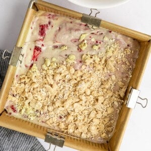 Square baking pan with strawberry batter topped with crumble next to a bowl of crumble on a white surface. A dark striped cloth is nearby.