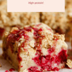 Close-up of a raspberry breakfast cake with crumb topping and visible red raspberry filling, placed on parchment paper. Text above reads "Raspberry Breakfast Cake - High-protein!.