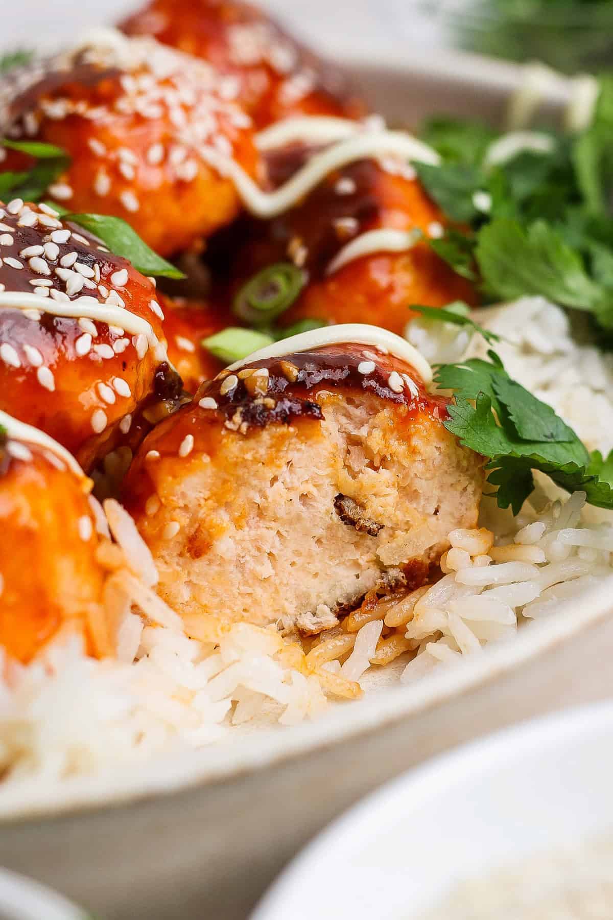 Close-up of rice topped with saucy chicken meatballs, garnished with sesame seeds and cilantro. A meatball is cut open, revealing its interior. Drizzled sauce is visible on top.