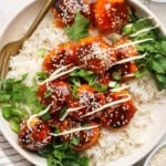 A dish of rice topped with saucy meatballs, drizzled with white sauce and garnished with sesame seeds and fresh herbs. A fork rests on the plate.