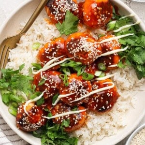 A dish of rice topped with saucy meatballs, drizzled with white sauce and garnished with sesame seeds and fresh herbs. A fork rests on the plate.