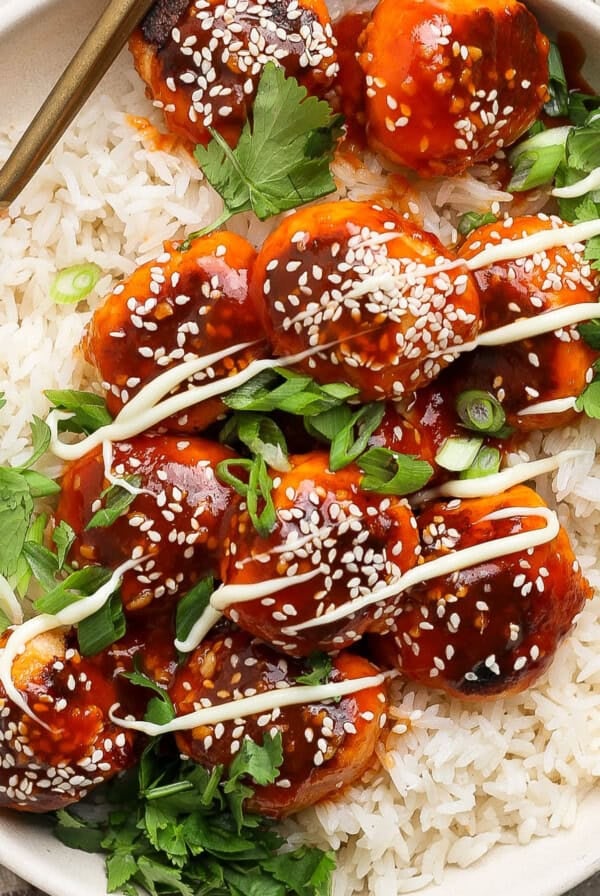 A dish of rice topped with saucy meatballs, drizzled with white sauce and garnished with sesame seeds and fresh herbs. A fork rests on the plate.