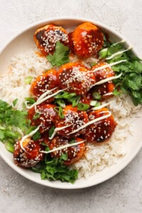A plate of rice topped with glazed meatballs, sesame seeds, green onions, drizzled sauce, and fresh cilantro.