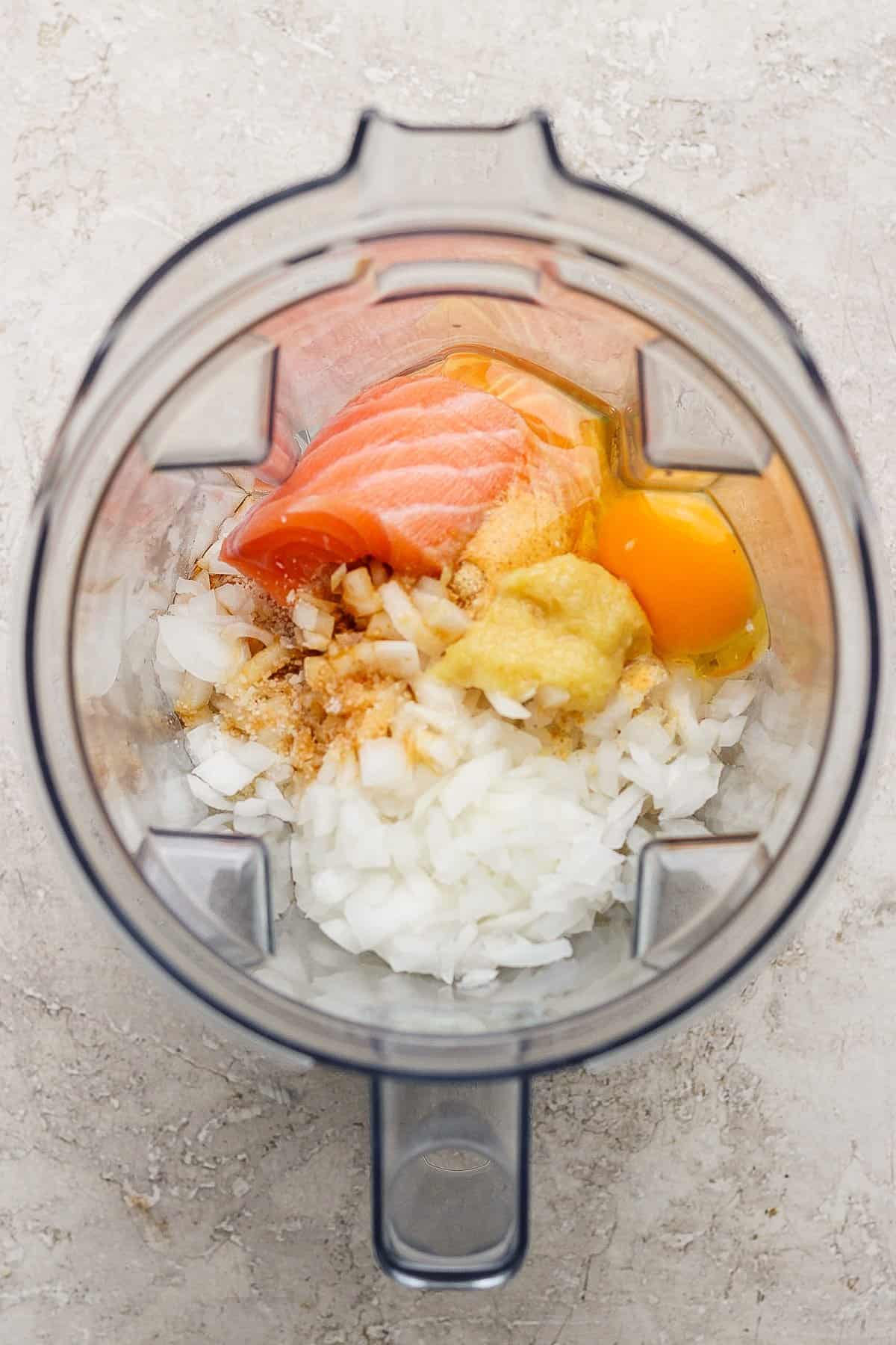 Top view of a blender containing chopped onion, raw salmon, an egg, mustard, and seasonings on a light textured surface.