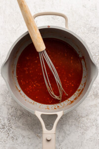 A whisk in a gray pan contains a thick red sauce with bits of seasoning.