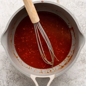 A whisk in a gray pan contains a thick red sauce with bits of seasoning.