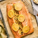 A stuffed salmon fillet crowned with herbs and spices, garnished with lemon slices, rests on parchment paper on a baking tray beside a spatula.