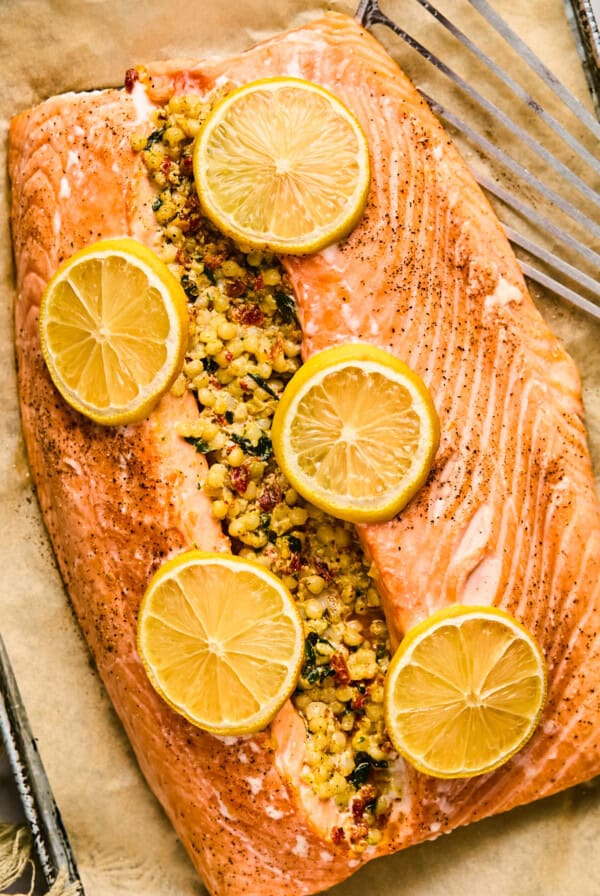 A stuffed salmon fillet crowned with herbs and spices, garnished with lemon slices, rests on parchment paper on a baking tray beside a spatula.