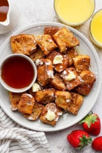 A plate of French toast bites with butter and powdered sugar, served with a small bowl of syrup. Two glasses of orange juice and fresh strawberries are on the side.