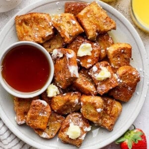 A plate of French toast bites with butter and powdered sugar, served with a small bowl of syrup. Two glasses of orange juice and fresh strawberries are on the side.