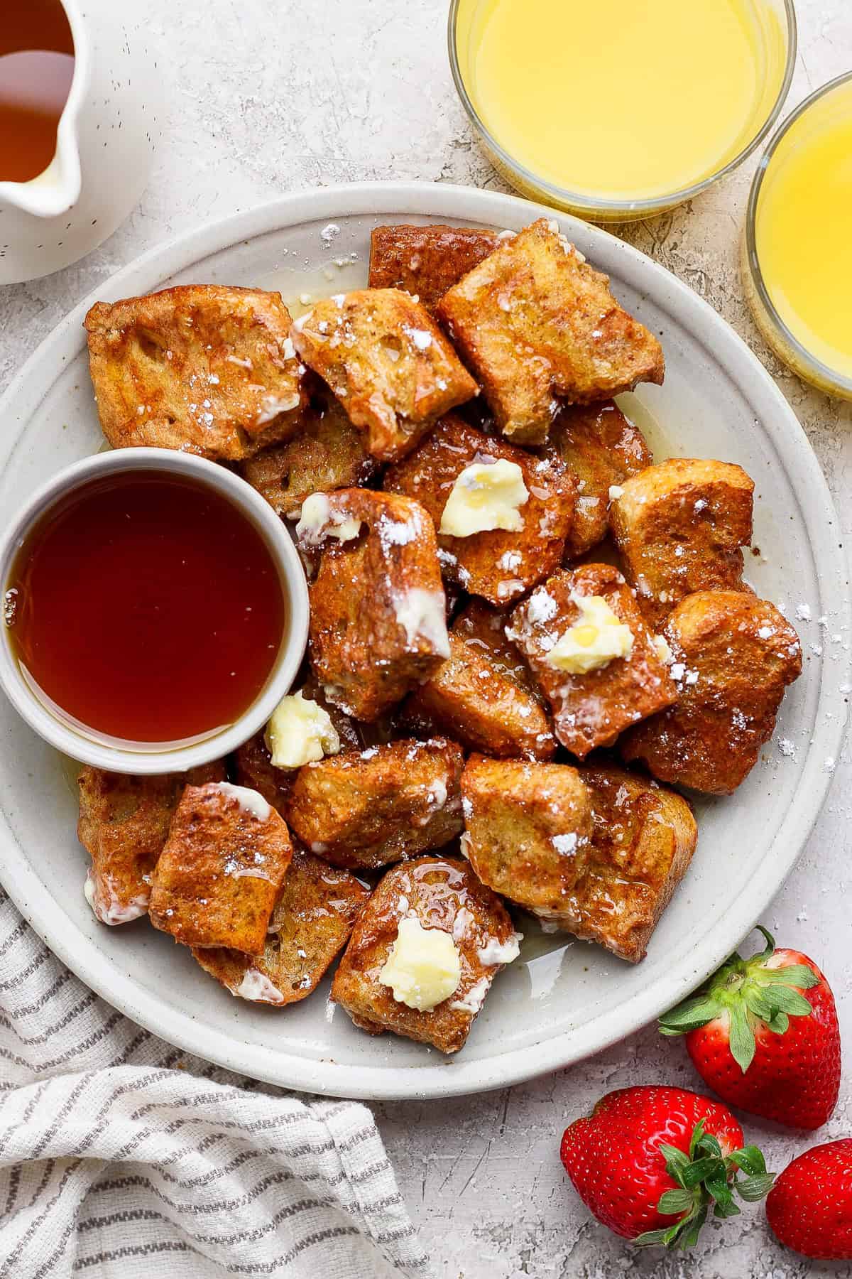 A plate of French toast bites with butter and powdered sugar, served with a small bowl of syrup. Two glasses of orange juice and fresh strawberries are on the side.