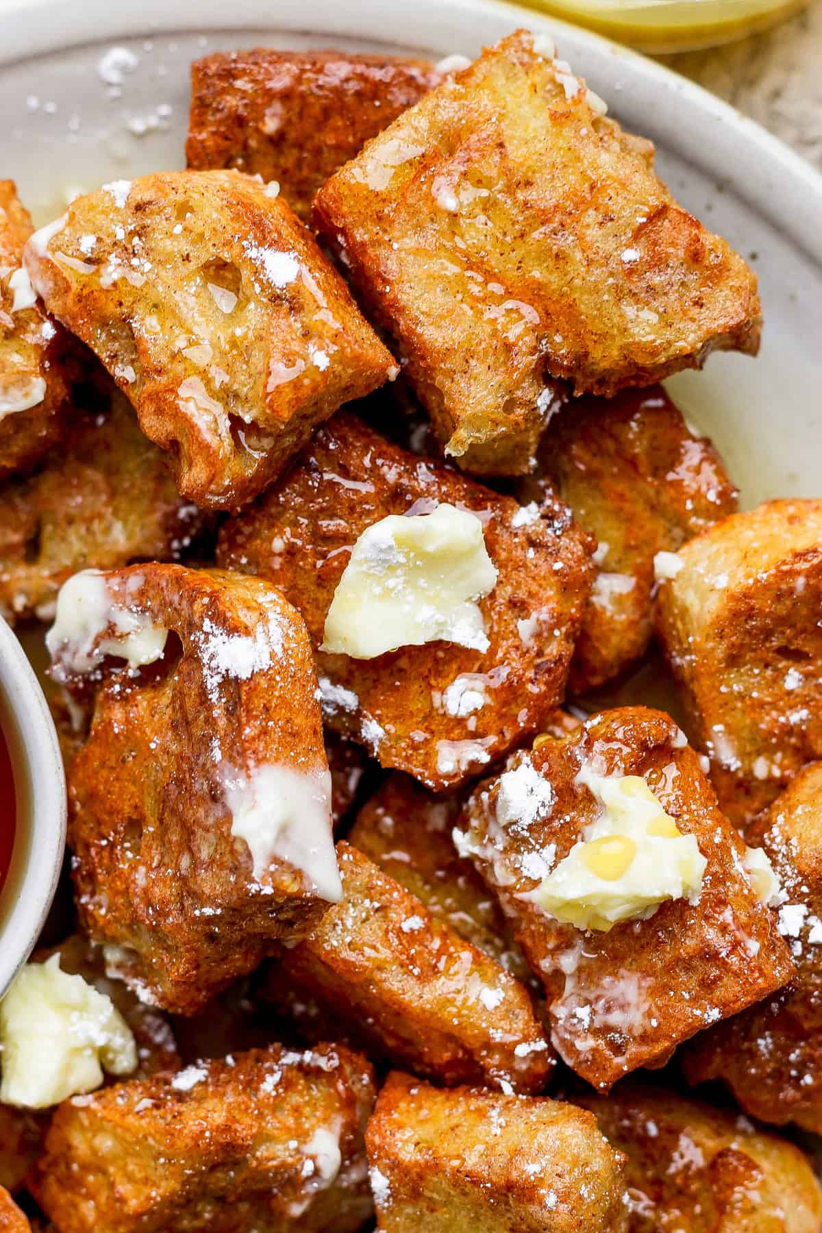 Close-up of French toast bites topped with melting butter and powdered sugar on a plate. A small bowl of syrup is visible in the corner.