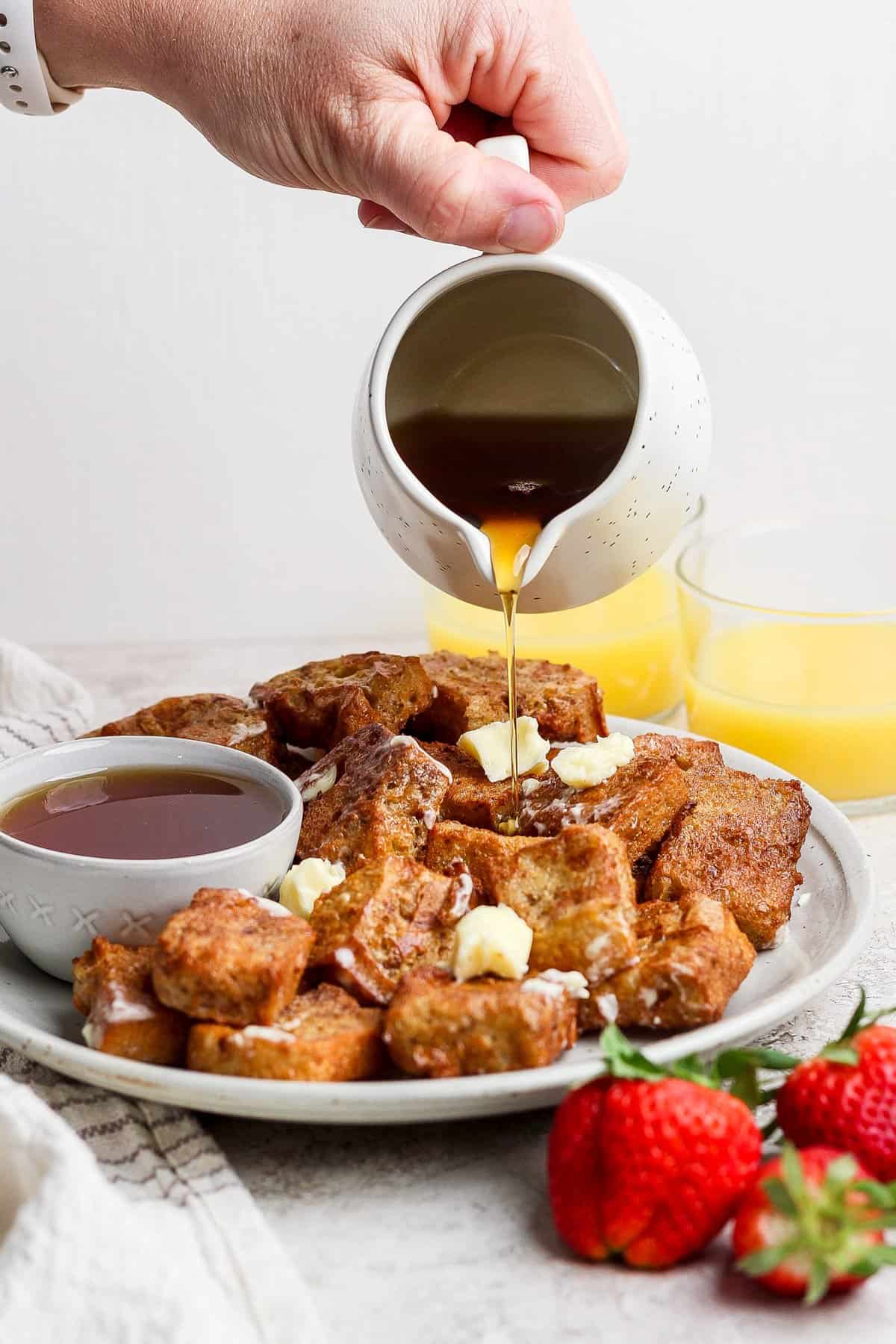 A hand pours syrup onto a plate of French toast cubes topped with butter. A bowl of syrup and strawberries are beside the plate. Two glasses of orange juice are in the background.
