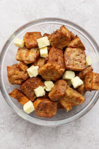 A glass bowl filled with croutons and small cubes of butter on a light gray textured background.