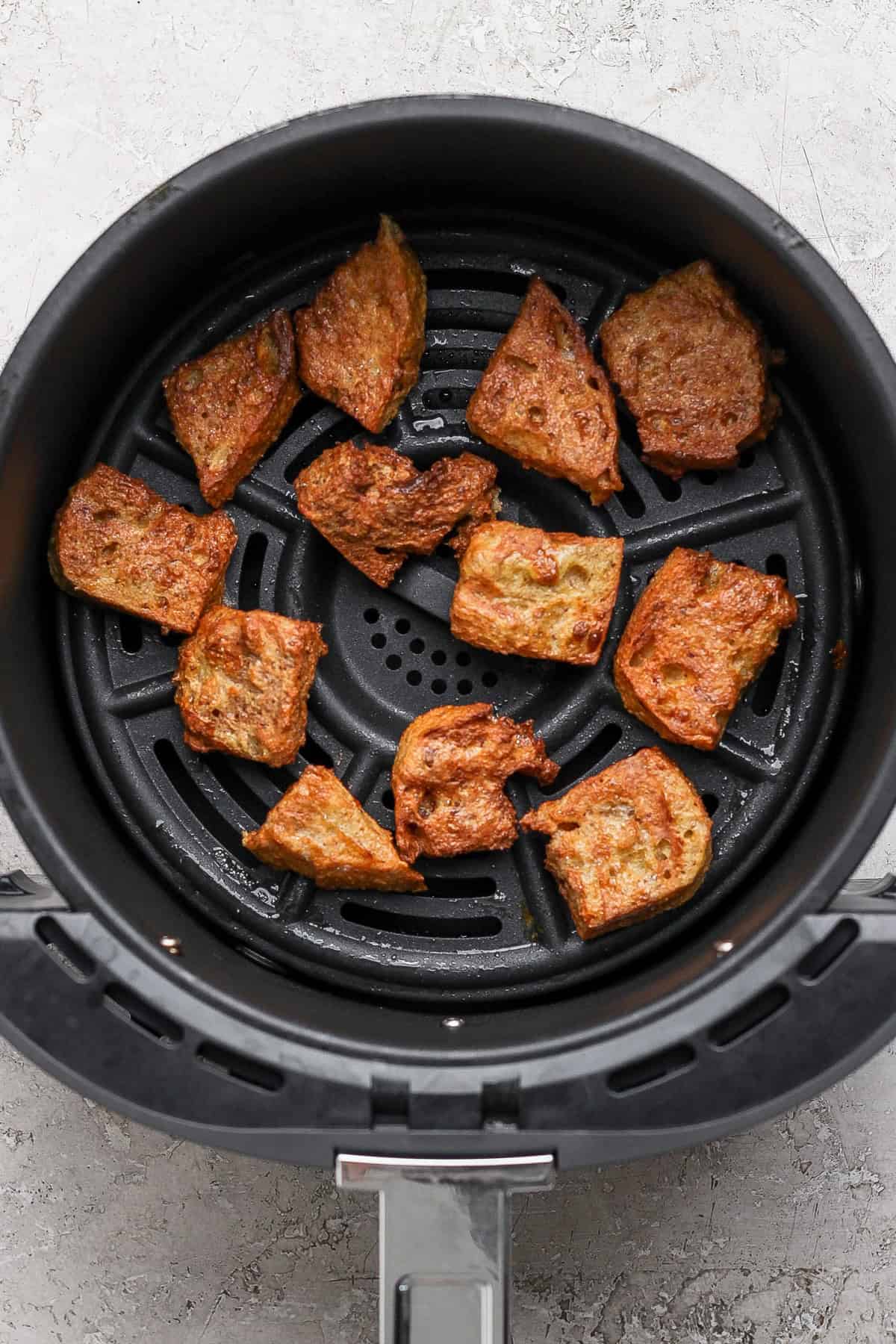 Crispy pieces of roasted bread sit in an air fryer basket on a light textured surface.