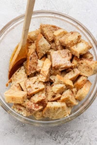 Bread cubes in a glass bowl with a wooden spoon, coated in a mixture, likely for a recipe. The bowl is placed on a light-colored, textured surface.