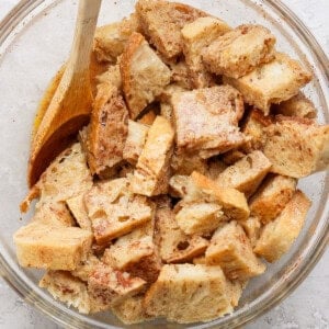 Bread cubes in a glass bowl with a wooden spoon, coated in a mixture, likely for a recipe. The bowl is placed on a light-colored, textured surface.