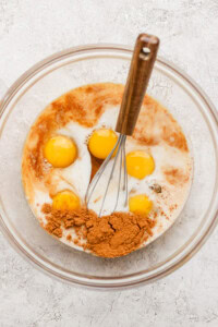 A mixing bowl contains eggs, cocoa powder, milk, and other liquid ingredients, with a whisk resting inside.