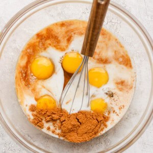 A mixing bowl contains eggs, cocoa powder, milk, and other liquid ingredients, with a whisk resting inside.