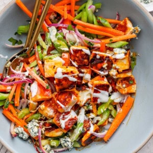 A salad with grilled chicken pieces, carrots, celery, red onion, blue cheese crumbles, drizzled with dressing, in a large bowl with two spoons.