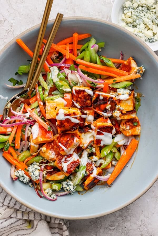 A salad with grilled chicken pieces, carrots, celery, red onion, blue cheese crumbles, drizzled with dressing, in a large bowl with two spoons.