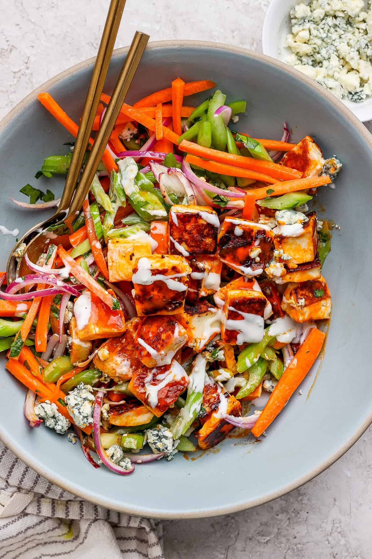 A salad with grilled chicken pieces, carrots, celery, red onion, blue cheese crumbles, drizzled with dressing, in a large bowl with two spoons.
