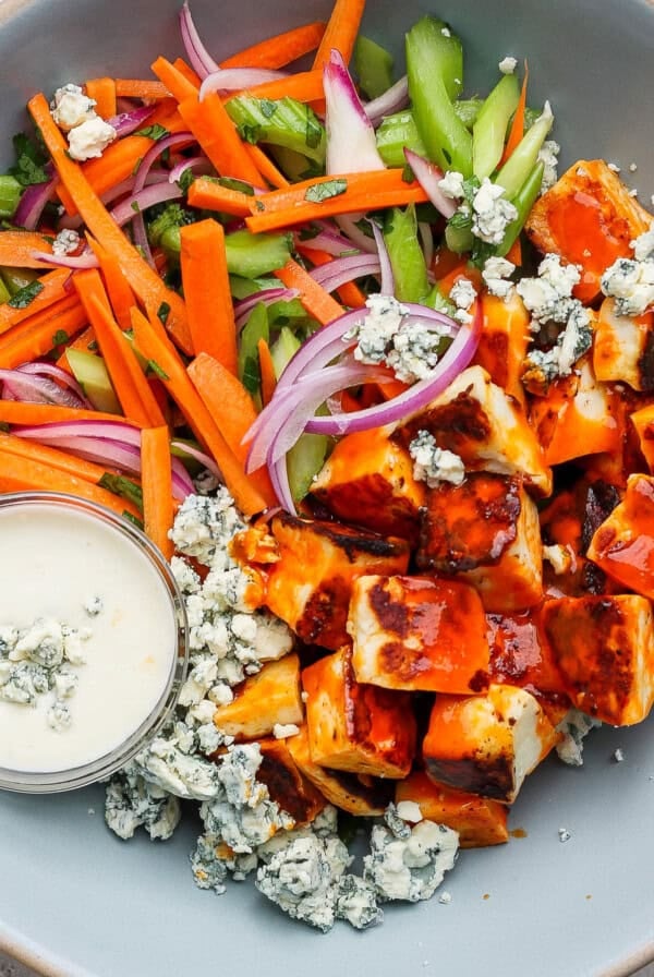 A bowl with diced carrots, celery, red onion, blue cheese crumbles, and buffalo sauce-coated tofu. A small cup of creamy dressing is on the side.