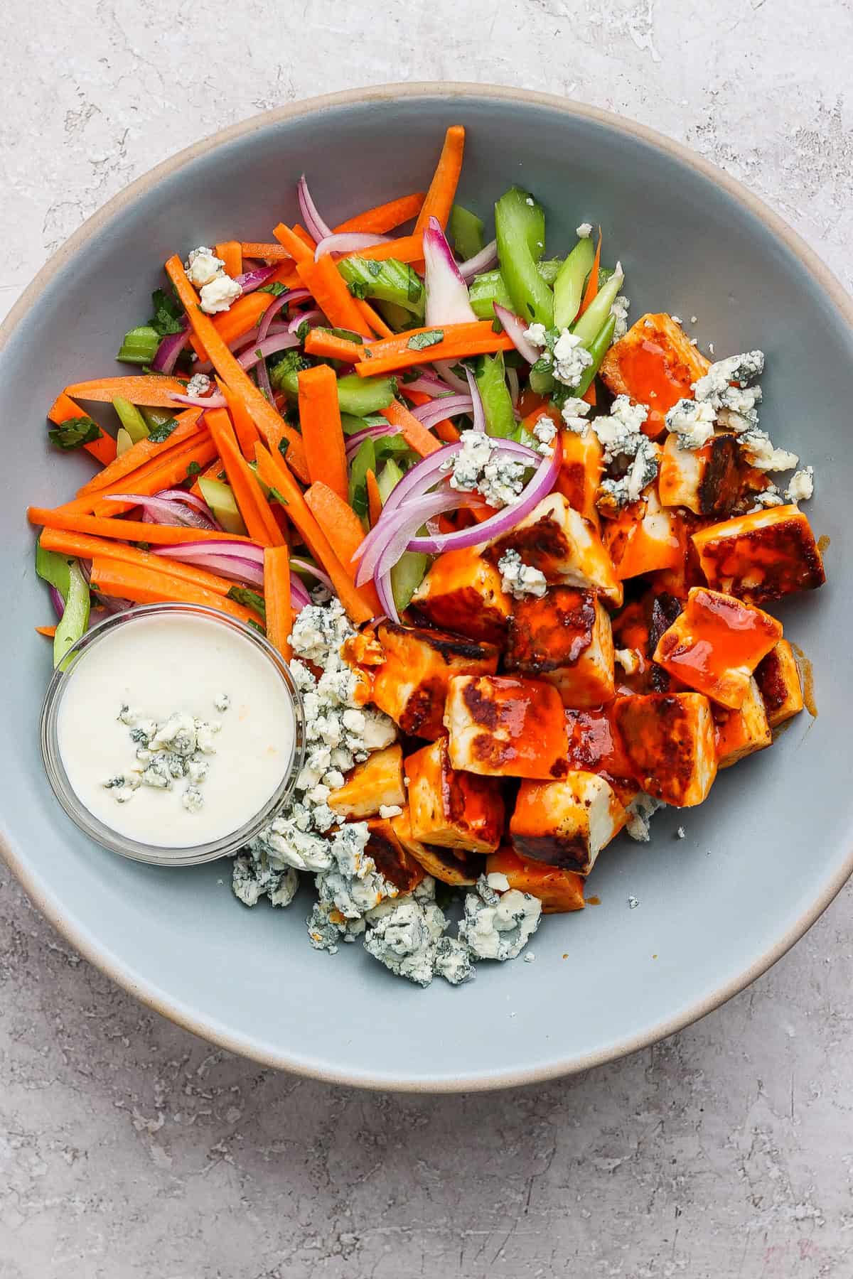 A salad bowl with cubed grilled chicken, crumbled blue cheese, carrot sticks, celery, red onion, and a small dish of creamy dressing on the side.