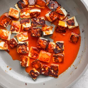 Pan-fried paneer cubes coated in a rich red sauce in a non-stick pan on a light gray surface.