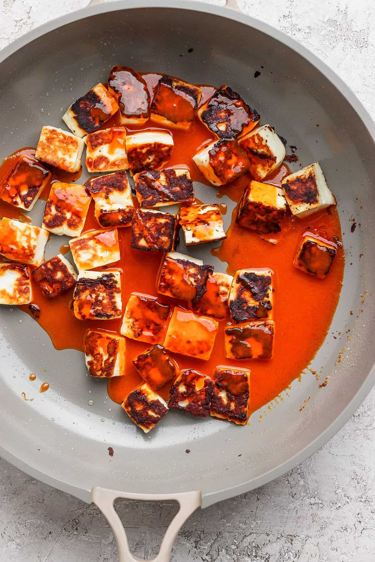 Pan-fried paneer cubes coated in a rich red sauce in a non-stick pan on a light gray surface.