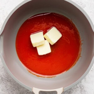 A grey pan contains a red liquid with three chunks of butter melting in it. The background is a light, textured surface.