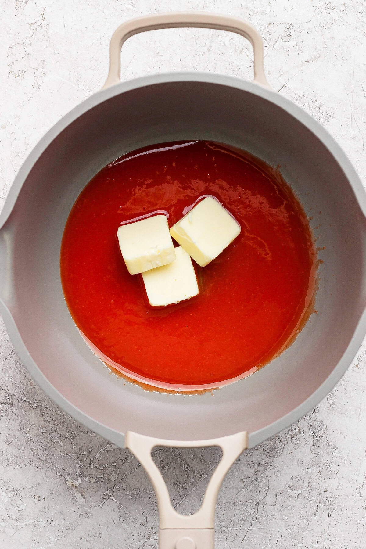 A grey pan contains a red liquid with three chunks of butter melting in it. The background is a light, textured surface.