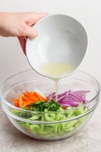 Person pouring a light dressing into a bowl of chopped vegetables including carrots, red onions, celery, and herbs.